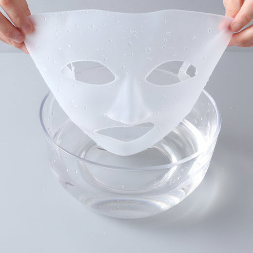 A reusable silicone face mask being washed, held up by two hands over a clear bowl of water